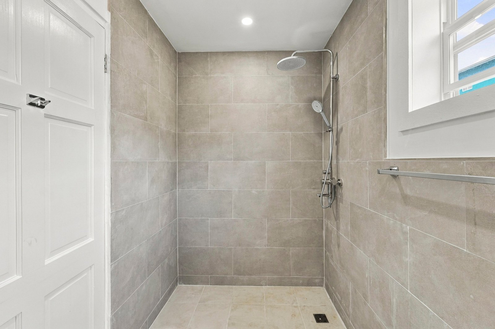 Grey-tiled shower area at Cinnamon Lot 120, with overhead and handheld shower fittings, a window and a floor drain.